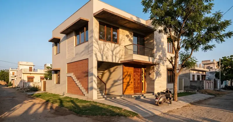 A two-storey west-facing house in Jaipur in late afternoon sun, with deep concrete sunshades over the windows, a terracotta jaali on the staircase wall and a neem tree shading the front. Illustrative image, made with AI.